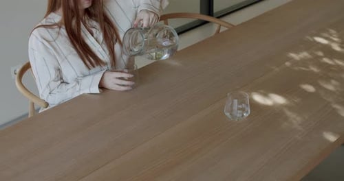Woman Pours Water into Glass on Wooden Table