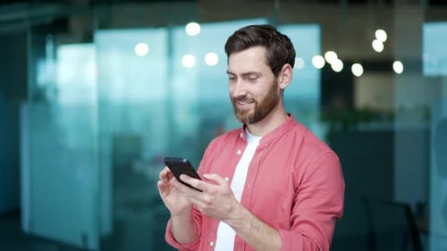 Lächelnder Geschäftsmann, Freiberufler, hält sein Smartphone in der Hand, scrollt und schaut soziale Medien im modernen Büro
