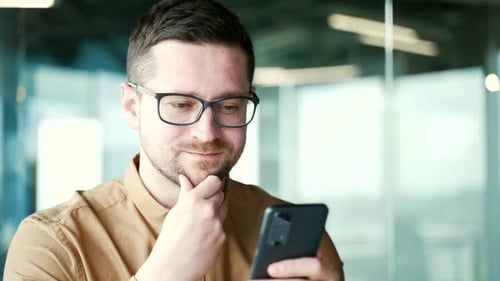 Close up. Thoughtful businessman in glasses is using smartphone at workplace in business office.