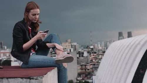 Young woman sitting on terrace and text messaging city in background