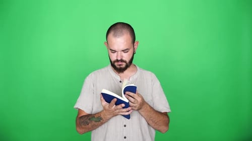 tattooed man flips through blue book in front of green curtain