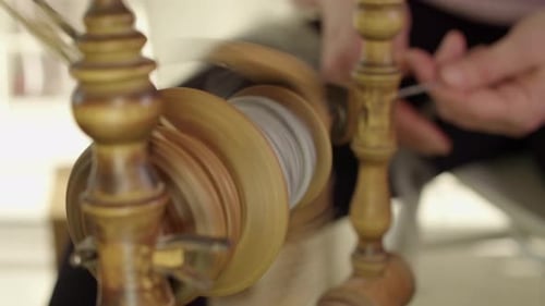 Spinning wheel bobbin foreground, defocused artisan hands background