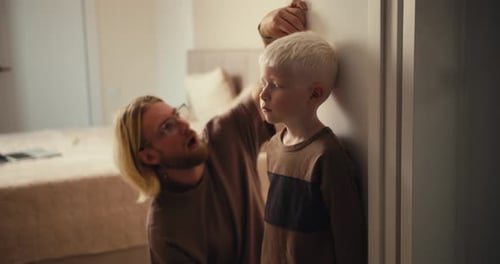 A Blond Car with Glasses and a Beard Measures the Height of His Little Albino Boy with White Hair