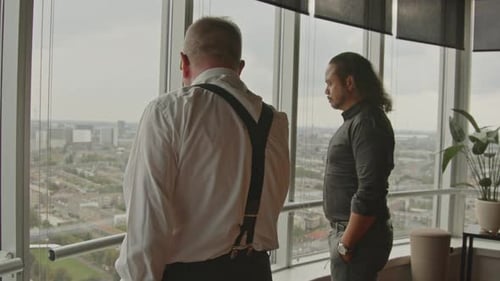 Two businessmen stand in front of an office window overlooking a city skyline and talk.