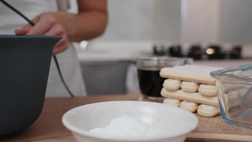The ingredients for the tiramisu cake are laid out on the table, while a woman is whipping the cream