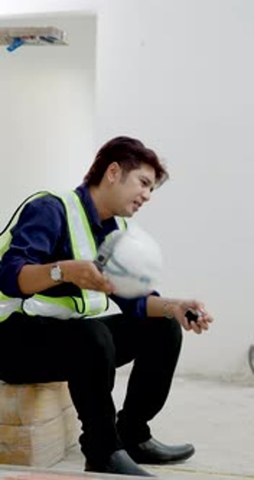 Asian male construction worker rests on brick ledge with helmet in hand, looking tired during