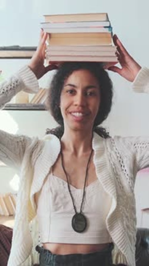 Young Optimistic African American Woman Student Puts Books on Head Sits on Sofa