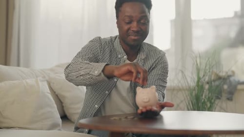 Porträt eines afrikanischen Mannes, der Geld in einer Münzbox spart und Münzen in ein Sparschwein steckt, junger erwachsener Mann