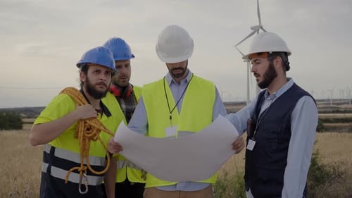 Group of Engineers Talking and Working with a Blueprint in an Agricultural Field with Wind Turbines