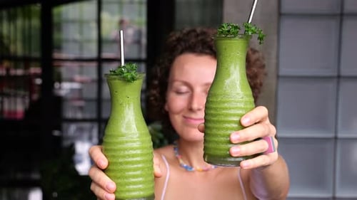 A Happy Woman Holds Out Two Bottles of Fresh Green Smoothie Filled with Vitamins and Energy for the