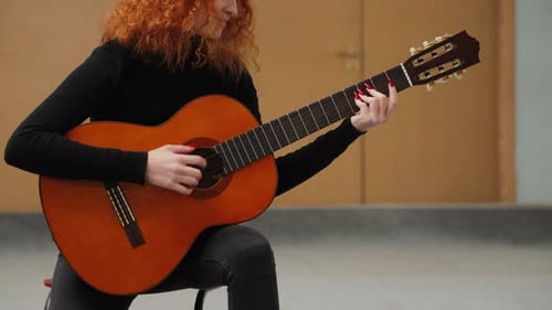 Close-up video of a woman playing chords on an acoustic guitar while sitting on a stool in a room.