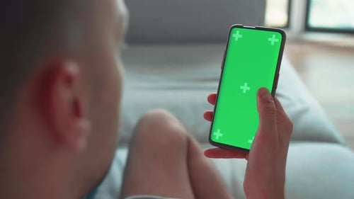 Young Caucasian Man at Home Lying on a Couch Using Smartphone Swiping with Green Mockup Screen Doing