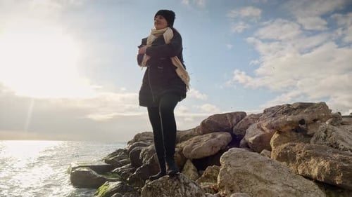 Young woman standing on the cliff, looking at the winter sunset over the sea