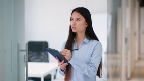 Young Asian Woman Reviewing Notes in an Office Setting While Taking Notes on a Clipboard
