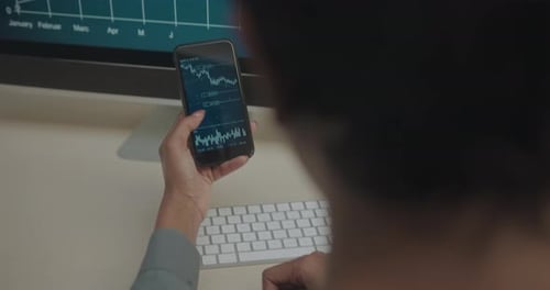 Female Stockbroker looking at Smart Phone Sitting at Desk in Office