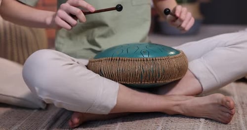 Woman playing steel tongue drum with mallets on floor at home, closeup