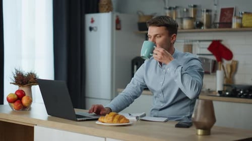 Young man works on laptop at home kitchen with coffee surfing the internet