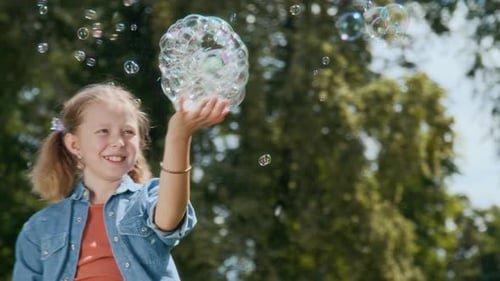 Joyful Pre-Teenage Girl Holding Soap Bubbles on Her Palm on Sunny Day Outdoors