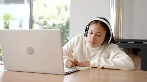 Wearing headphones, girl studying at home with laptop and notebook, copy space