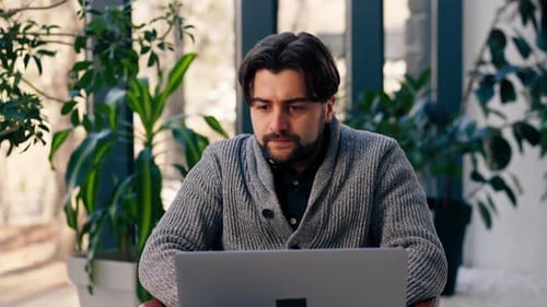 of Young Guy Office Worker Against the Background of a Window Sitting on a Chair Working at a Laptop