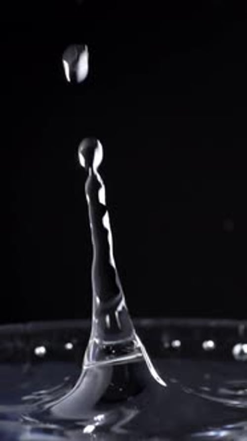 Vertical Shot Of Water Droplets Splashing On Water Surface In Glass With Black Background, Close-Up