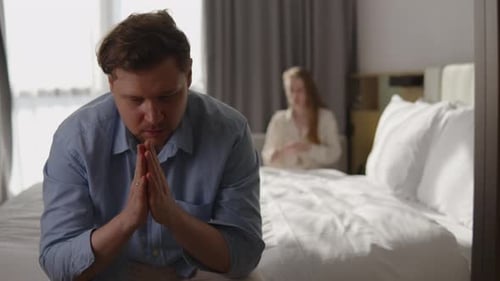 Worried Man Sitting on Bed Feeling Stressed and Insecure with Woman in Background Looking