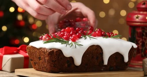 Woman putting cranberries onto tasty Christmas cake with icing and rosemary at wooden table, closeup