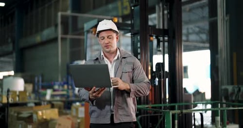 caucasian male warehouse employee using laptop while managing supply chain operation near forklift