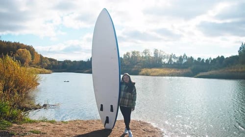 Young Attractive Woman Stands Before Large Buoyant Surfboard Against the Backdrop of Serene Pond on