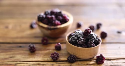 Ripe frozen blackberries in bowls on wooden table, closeup. Camera moving forward