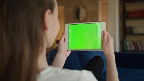 Back View of Young Female Holding Horizontal Tablet with Green Screen Sitting in Room
