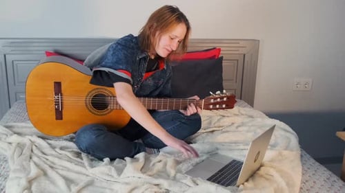 Young Woman Plays Guitar on Bed with Laptop