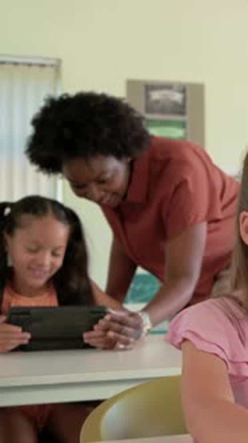 Vertical video: Leaning teacher in rust top teaching student in class after tap, pointing at tablet