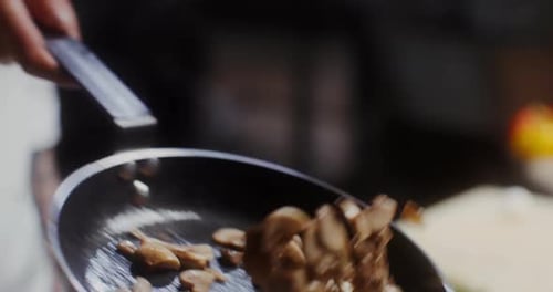 A man in a chef's uniform mixes mushrooms by tossing them up from the frying pan, close-up