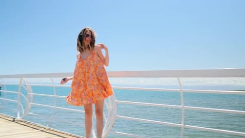 Young woman blonde in flying orange dress dancing on the pier by the sea listening music in headphon