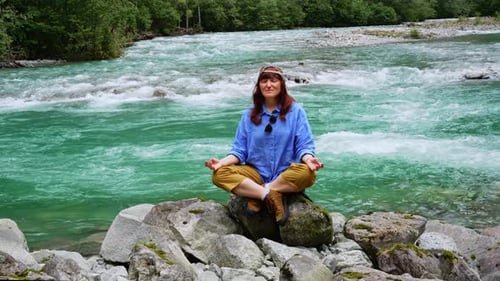 Girl Wearing a Blue Shirt Sits on a Large Stone and Meditates Near a Waterfall Next to a Mountain