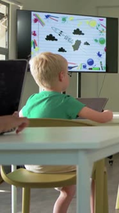 Vertical video: Watching blond boy in green tee lifting tablet after monitor art shows in classroom