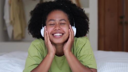 Jolie adolescente noire aux cheveux bouclés avec des écouteurs sur le lit rit et sourit avec ses dents tout en