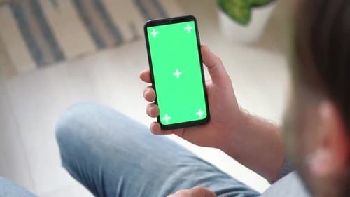 Young Man Sitting at Home Holding Smartphone Green Mockup Screen in Hand Male Person Using Chroma