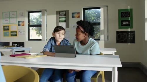 Two preteen classmates watching tablet in class reacting to teacher appearing, working with notes