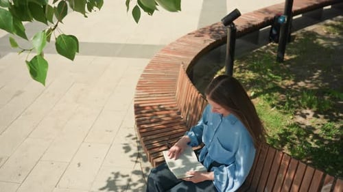 White Woman Seated on Curved Bench in Sunny Plaza Reading and Jotting Notes Aerial View Shows