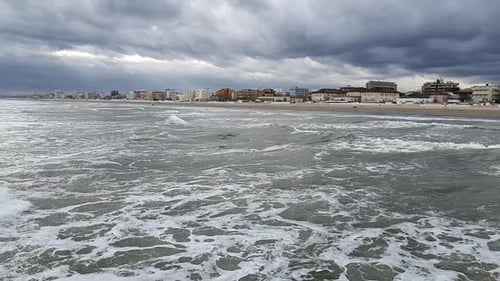 Rough sea waves rushing against beach city in stormy weather. Bad weather on the shore. Buildings ne