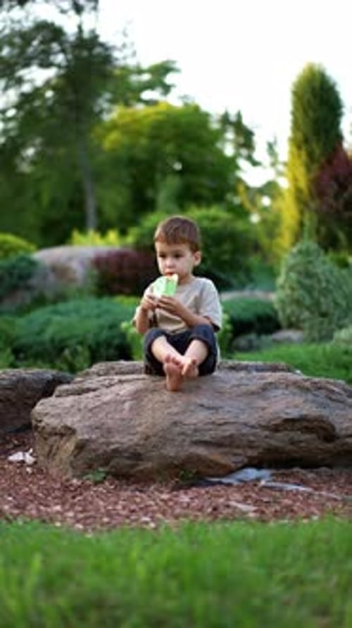 Adorable Caucasian toddler sitting on the stone in the park. Baby boy eating from a doy pack.