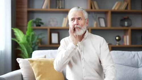 Upset elderly senior man with gray hair suffering from toothache sitting on the sofa in the living