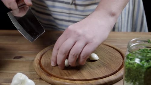 Chef cutting stems of three cloves of garlic, uses kitchen ax knife on round wooden board. Garlic, a
