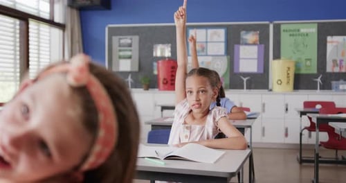 Happy diverse schoolgirls raising hands at desks in elementary school class, slow motion