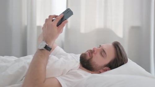 Side View of Young Adult Man using Phone in Bed