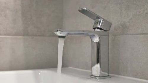A close-up shot of a modern chrome bathroom faucet with water flowing into a white sink. A hand is