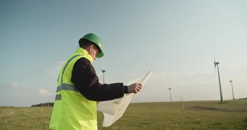 An engineer in a reflective vest stands with design plans at a wind farm.