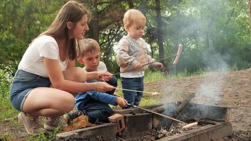 Young mother with two sons cooking sausages on bonfire in camp